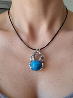 Halsband av rostfritt stål med drakklo blå - Ett halsband med ett hänge som föreställer en klo som håller en blåskimrande kula. Halsbandet har en svart rem och ett justerbart silverfärgat spänne. Berlock 3,7 cm hög.