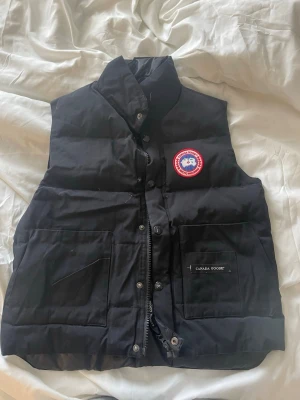 Svart väst från Canada Goose - Äkta svart dunväst från Canada Goose med hög krage och två stora fickor framtill. Västen har Canada Goose-logga på bröstet och dragkedja med knappar. Lite skavanker på knappar, men syns ej. Modell: 4565MR. Storlek: XS/P.