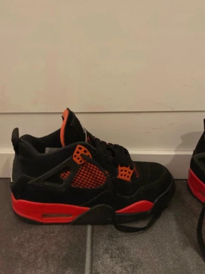 Svarta och röda Air Jordan 4 sneakers - Snygga Air Jordan 4 sneakers i svart och rött med röda detaljer. Skorna har klassisk meshpanel på sidorna, Jumpman-logga på hälen och vadderad plös. Ovandelen är i mocka och textil, sulan är i gummi. Perfekta för dig som gillar streetwear och basketstil. ÄKTA ingen kopia