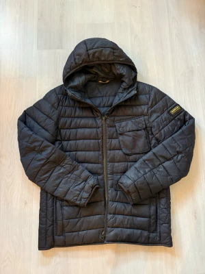 Svart quiltad pufferjacka Barbour - Svart quiltad pufferjacka från Barbour International med huva och dragkedja framtill. Jackan har en bröstficka med knapp och en patch med Barbour-logga på ärmen. Perfekt för kyliga dagar och riktigt snygg streetstil.