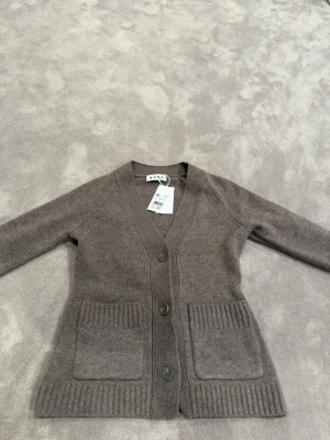 Brun kofta från Wera med fickor - Mysig brun kofta från Wera med tre knappar framtill och två stora ribbstickade fickor. Koftan har v-ringning och är tillverkad i mjukt stickat material. Perfekt för lager-på-lager och enkel att matcha med olika outfits.