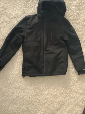 Svart vindjacka med huva från Didriksons - Svart vindjacka från Didriksons med huva och dragkedja framtill. Jackan har två bröstfickor med dragkedja och reflexdetaljer på ärm och rygg. Perfekt för blåsiga dagar och enkel att matcha med olika outfits.