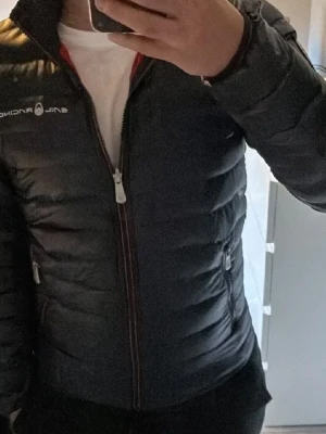 Sail Racing jacka - Säljer en svart dunjacka från Sail Racing med vit dragkedja och logga på bröstet. Jackan har quiltad design, två sidofickor med dragkedja och röd insida. Perfekt för kalla vinterdagar och riktigt snygg till streetwear-stilen.