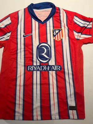 Atlético Madrid matchtröja Nike röd - Snygg Atlético Madrid matchtröja från Nike i rött med vita och blå vertikala ränder. Tröjan har klubbmärke på bröstet, sponsorlogga 'Riyadh Air' och blå krage. Tillverkad i lätt och ventilerande funktionsmaterial, perfekt för fotbollsträning eller match.