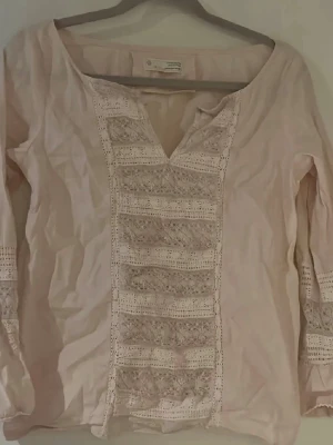 Ljusrosa blus från Odd Molly - Supersöt ljusrosa blus från Odd Molly med virkade spetsdetaljer framtill och på ärmarna. Blusen har en v-ringning och är tillverkad i ett mjukt tyg som känns skönt mot huden. Perfekt för dig som gillar boho-vibbar och romantiska detaljer.