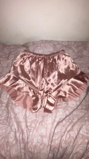 Rosa satinshorts med volangkant - Supersöta pyjamasshorts i glansigt rosa satin med bred volangkant nedtill och elastisk midja. Perfekta för mysiga kvällar hemma och ger en lyxig känsla.