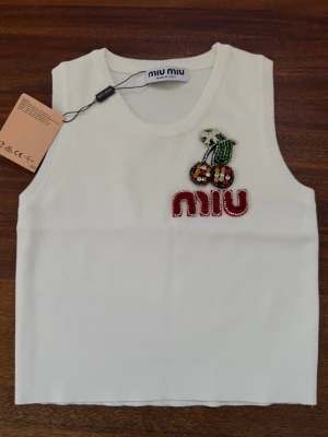  Miu Miu topp - Superfin vit linnetopp från Miu Miu med pärlbroderade körsbär och logga framtill. Toppen är ärmlös och har en klassisk rund halsringning. Perfekt för att lyfta vilken outfit som helst med en söt detalj.