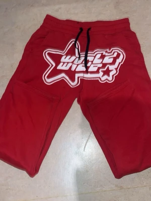 Röda byxor med tryck från FSBN - Säljer ett par röda byxor från FSBN med stort vitt tryck och stjärndetalj framtill. Shortsen har snörning i midjan och är tillverkade i mjuk bomullsmix. Perfekta för en avslappnad och sportig stil.