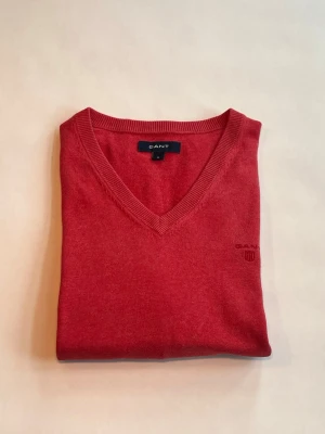 Röd V-ringad tröja från GANT - Snygg röd V-ringad tröja från GANT med diskret broderad logga på bröstet. Tillverkad i mjukt material och har ribbade muddar vid ärmslut och nederkant. Perfekt för dig som vill ha en klassisk och stilren look. Har ett hål i ena armhålan. 