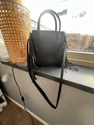 Svart handväska med fransar Don Donna - Snygg svart handväska från Don Donna i skinnimitation med långa fransar på sidorna. Väskan har både handtag och en lång axelrem så du kan bära den på flera sätt. Stilren design med guldfärgad logga upptill. Perfekt accessoar för att lyfta din outfit.