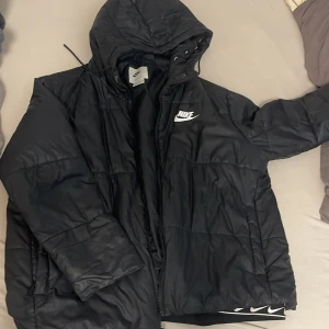 Svart Nike pufferjacka Therma-Fit XL - Svart pufferjacka från Nike i modellen Therma-Fit. Jackan har huva, dragkedja och coola Nike-loggor längs insidan. Oversized passform och tillverkad i syntetmaterial. Perfekt för kalla dagar när du vill hålla stilen.