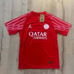 Röd PSG x Jordan fotbollströja - Säljer en röd PSG x Jordan fotbollströja med vit logga och Qatar Airways-tryck på bröstet. Tröjan har korta ärmar med vita grafiska linjer och är gjord i lätt, ventilerande Dri-Fit material. Perfekt för dig som vill sticka ut på planen.