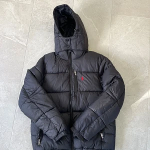 Svart pufferjacka Polo Ralph Lauren - Svart pufferjacka från Polo Ralph Lauren med röd broderad logga på bröstet. Jackan har huva, flera dragkedjor och quiltad design. Perfekt för kalla dagar och har en clean, sportig look.