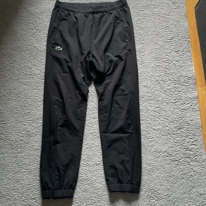 Svarta tapered träningsbyxor Lacoste - Svarta träningsbyxor från Lacoste Sport med tapered fit och elastisk midja. Byxorna har snörning, dragkedjor vid fickorna och klassisk Lacoste-logga på benet. Materialet är lätt och syntetiskt, perfekt för sportiga aktiviteter.