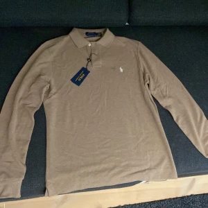 Brun långärmad pikétröja Polo Ralph Lauren - Snygg brun långärmad pikétröja från Polo Ralph Lauren i custom slim fit. Tröjan har klassisk krage, knappar vid halsen och den ikoniska broderade loggan på bröstet. Tillverkad i mjuk bomull som känns skön mot huden. Perfekt för dig som gillar stilrena och tidlösa plagg.