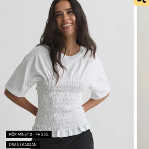 Vit smockad topp med volang - Vit topp med smockad midja och volangkant nertill. T-shirten har korta ärmar och rund halsringning. Perfekt för en fräsch och stilren look.