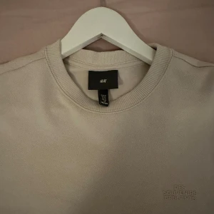 Beige sweatshirt från H&M - Säljer en stilren beige sweatshirt från H&M med broderad text 'DES SOUVENIRS BRULANTS' på bröstet. Tröjan har rund halsringning, långa ärmar och ribbade muddar. Perfekt för en chill och avslappnad look.