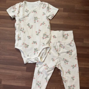 Set med smultron - Mjukt set med omlottsbody och byxor från Lindex med smultronmönster i storlek 68 (4-6mån). Oanvänd och i nyskick. Däremot lyckades plaggen hamna i en grövre tvätt och loggan har blekts bort en aning, men absolut ingenting annat tog skada.