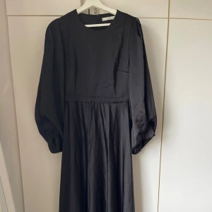 Svart långklänning med puffärmar - Snygg svart långklänning från Lemoniade med rund halsringning och puffiga långärmar. Klänningen har en markerad midja och ett mjukt, flytande tyg som ger en elegant siluett. Dragkedja i ryggen för enkel påklädning. Perfekt för dig som gillar stilrena och klassiska looks.