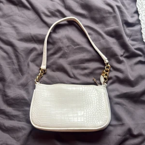 Vit axelväska med krokodilmönster - Snygg vit axelväska med krokodilmönstrad yta och guldfärgade kedjedetaljer på axelremmen. Väskan har en praktisk dragkedja upptill och en stilren, rektangulär form som gör den enkel att matcha med olika outfits. Perfekt storlek för det viktigaste.