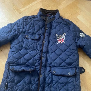 Blå quiltad jacka GeoNorway L - Snygg marinblå quiltad jacka från GeoNorway Royal Club med broderad logga på bröstet, stora fickor med knappar och förstärkta armbågar i manchester. Jackan har hög krage, tryckknappar och coola detaljer som ger en sportig och klassisk look.