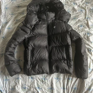 Svart pufferjacka från Calvin Klein - Svart pufferjacka från Calvin Klein. Jackan har huva, två dragkedjeförsedda fickor och quiltad design. Perfekt för kalla dagar och har en clean, modern look. Trycket har åkt av lite(bild 3,4) men annars så är den i super fint skick och jag tycker inte att man märker det särskilt mycket 