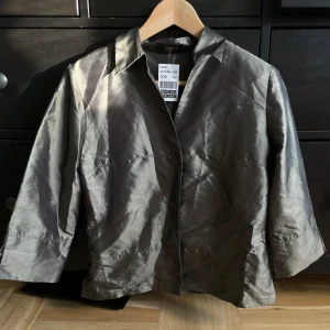 Silvrig/Grön kavajjacka från Flash - Silvrig kavajjacka från Flash med glansig finish och klassisk krage. Jackan har trekvartsärmar, knäppning framtill och en rak, stilren passform. Perfekt för dig som vill sticka ut med en metallic look.