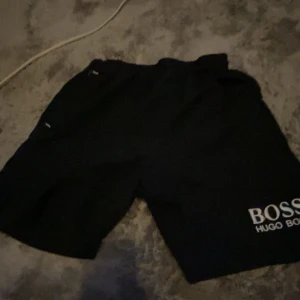 Svarta shorts från Hugo Boss - Svarta shorts från Hugo Boss med vit broderad logga på benet. De har vit snörning i midjan och meshfoder på insidan. Perfekta för sommaren och har en clean, sportig vibe.