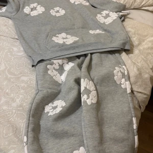 Grå hoodie från Denim Tears med blommor - Säljer en grå hoodie från Denim Tears med vita blommiga tryck över hela tröjan. Hoodien har en klassisk huva och magficka, och är fodrad med mjuk fleece på insidan. Perfekt för dig som gillar streetwear och vill sticka ut med snygga detaljer.