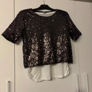 Svart paljettblus med vit underdel - Säljer en svart blus med glittriga paljetter och korta ärmar. Toppen har en vit, mjuk underdel som sticker ut nedtill och ger en cool lager-på-lager look. Perfekt för dig som vill sticka ut med lite extra bling och stil.