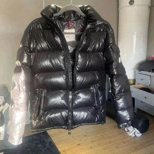  Moncler Maya  - Säljer en svart pufferjacka från Moncler med glansig finish och klassisk logga på ärmen. Jackan har hög krage, två dragkedjefickor fram och quiltad design. Perfekt för kalla dagar och riktigt snygg till jeans.