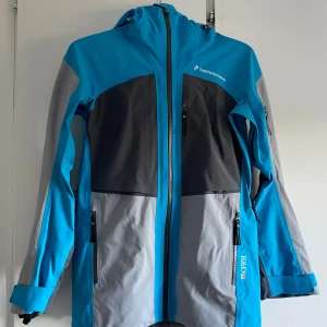 Blå och grå skaljacka Peak Performance GoreTex - Snygg skaljacka från Peak Performance i blått, grått och svart. Jackan har huva, flera dragkedjeförsedda fickor och justerbara ärmslut. Perfekt för utomhusaktiviteter och har en sportig look med logga på bröstet och ärmen. GoreTex. 