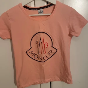 Rosa Moncler t-shirt med stor logga - Snygg rosa t-shirt  med stor logga framtill i svart och rött. Klassisk rund halsringning och korta ärmar. Perfekt för dig som gillar exklusiva märken och vill sticka ut med en cool detalj på bröstet.