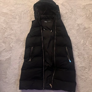 Svart dunväst från Zara - Lång svart dunväst från Zara med huva och dragkedja framtill. Västen har två fickor med dragkedjor och justerbar dragsko vid huvan. Perfekt för hösten, vår och vinter