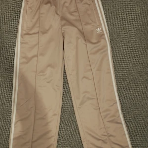 Beige Adidas firebird L - Snygga beige Adidas byxor med klassiska vita ränder längs sidorna och broderad logga framtill. Byxorna har dragkedja vid benslut, elastisk midja och är tillverkade i glansig polyester. Perfekta för en sportig och avslappnad stil.
