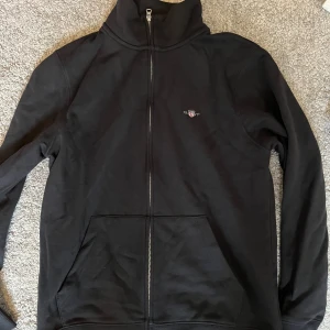 Svart ziptröja från GANT med logga - Svart ziptröja från GANT med broderad logga på bröstet och snygga ränder i kragen. Tröjan har lång ärm, två fickor fram och hel dragkedja. Perfekt för dig som gillar klassisk stil med sportig touch.