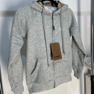 Grå hoodie från Burberry med dragkedja - grå hoodie från Burberry med klassisk logga broderad på bröstet, Perfekt för en chill och stilren look. Är i nyskick 