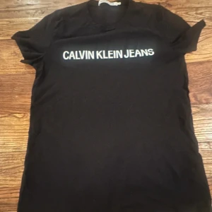 Svart t-shirt från Calvin Klein Jeans - Svart t-shirt från Calvin Klein Jeans med vit logotyp tryckt över bröstet. Klassisk rund hals och korta ärmar. Tillverkad i mjuk bomull som känns skön mot huden. Perfekt för dig som gillar stilrena och enkla plagg med tydlig branding.