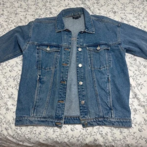  blå jeansjacka - Säljer en klassisk blå jeansjacka från Monki med raka linjer, två bröstfickor med knappar och silverfärgade knappar framtill. Jackan har en tidlös design och är perfekt för lager-på-lager-stil. Normal passform och långärmad.