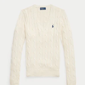 Vit kabelstickad tröja Polo Ralph Lauren. - Vit kabelstickad tröja från Ralph Lauren med v-ringning och marinblå logga på bröstet. Tröjan har lång ärm och ribbade muddar vid ärmslut och nederkant.