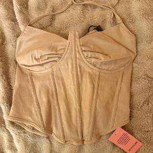 Beige korsett från PrettyLittleThing - Säljer en beige korsett från PrettyLittleThing med snygga sömmar och formade kupor. Toppen har justerbara axelband och dragkedja i ryggen för enkel på- och avtagning. Materialet är mjukt och har en lätt satin-finish som ger en lyxig känsla. Storlek (12), M passar även L