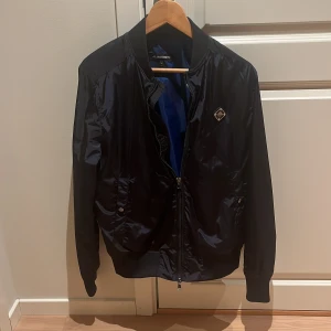 Svart bomberjacka från J.Lindeberg - Snygg svart bomberjacka från J.Lindeberg med glansig finish och klassisk passform. Jackan har dragkedja framtill, ribbade muddar och två fickor med tryckknapp. Perfekt för dig som vill ha en stilren och modern look. Helt nytt skick använd 2 gånger