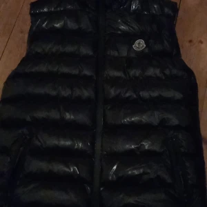 Svart dunväst från Moncler - Säljer en svart, glansig dunväst från Moncler med klassisk logga på bröstet. Västen har quiltad design, dragkedja framtill och två fickor. Perfekt för lager-på-lager och ger en clean streetstyle-vibe.