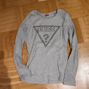 Grå ribbad långärmad tröja Guess XS - Supersnygg grå ribbad långärmad tröja från Guess med ikoniskt logotryck i silver framtill. Tröjan är i mjuk bomullsblandning och har en slim passform som sitter snyggt på kroppen. Perfekt till jeans eller kjol för en chill men trendig look.