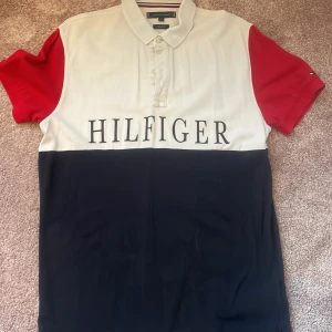 Tommy Hilfiger färgblockad piké M - Snygg pikétröja från Tommy Hilfiger i storlek M med färgblockad design i vitt, rött och mörkblått. Stor Hilfiger-logga framtill, klassisk krage och knappar. Regular fit och korta ärmar. Perfekt för dig som gillar stilren streetwear.