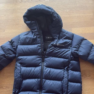 Marinblå pufferjacka från Sail Racing - Säljer en marinblå pufferjacka från Sail Racing med huva och dragkedja. Jackan är vadderad med breda kanaler och har en stilren logga på bröstet. Perfekt för kalla dagar när du vill hålla värmen och ändå se snygg ut. Har inga märken eller något.