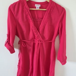 Rosa omlottklänning från Motherhood - Mamma blus. Snygg rosa omlottklänning från Motherhood Maternity med trekvartsärm och knytband i midjan. Klänningen har v-ringad hals och fina veckade detaljer längs urringningen. Perfekt för dig som vill ha en bekväm och stilren look.