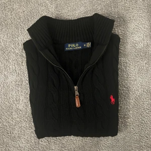 Cable Knit Half Zip Polo Ralph Lauren - Svart kabelstickad tröja från Polo Ralph Lauren med halv dragkedja och ribbad krage. Klassisk röd logobrodering på bröstet. Tröjan har långärmad passform och är tillverkad i mjukt stickat material som ger en snygg och tidlös look. Har använt den ett fåtal gånger men den är i fortfarande jätte bra skick.
