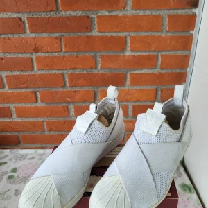 Vita Adidas Superstar Slip-On sneakers - Säljer ett par vita Adidas Superstar Slip-On sneakers med elastiska band över foten och meshdetaljer på sidorna. Skorna har klassisk snäcktå, dragflikar både fram och bak samt Adidas-logga på plösen. Perfekta för dig som gillar stilren och sportig look.