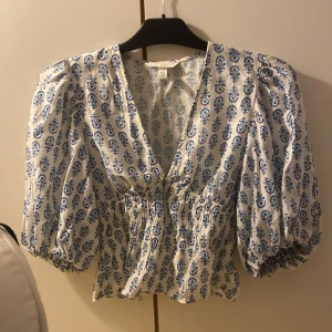 Vit och blå blus med puffärm från H&M - Säljer en vit blus från H&M i storlek XS med blått blommigt mönster. Blusen har puffiga korta ärmar, v-ringning och smockad midja som ger en snygg siluett. Perfekt för dig som gillar romantiska detaljer och vill sticka ut med en fräsch look. Passar folk med S också 